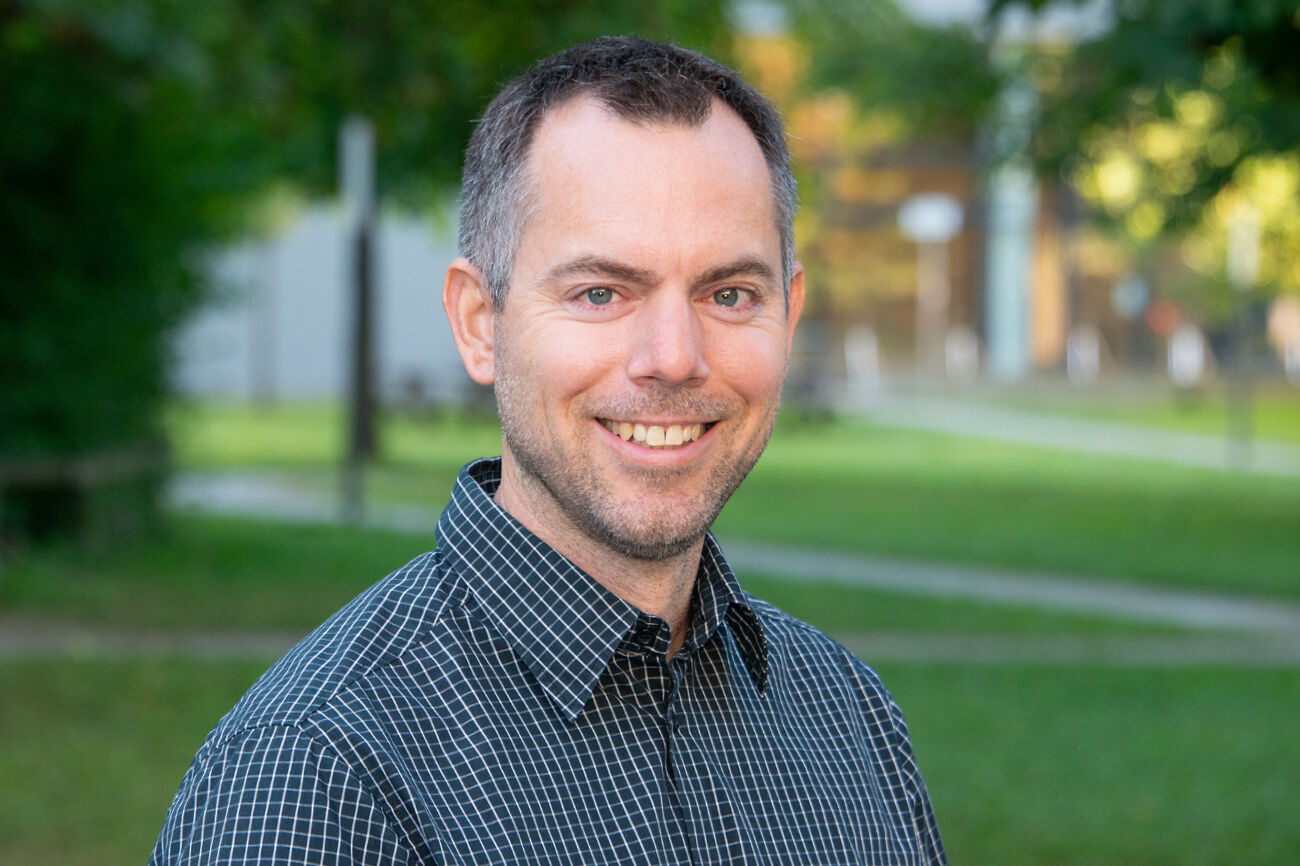 Mann in einem hellblauen Hemd, lächelnd im Freien vor grünem, unscharfem Hintergrund.