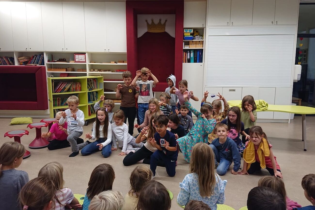 Kinder sitzen um einen Tisch herum und hören zwei Erwachsenen zu, die den Umgang mit Kräutern demonstrieren.