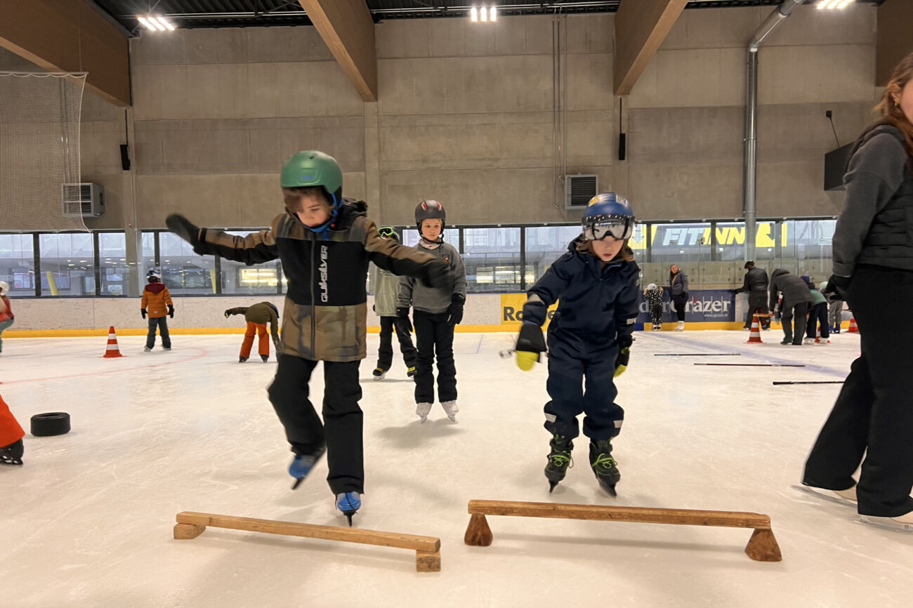 Zwei Buben probieren über ein Holzhindernis am Eis zu springen.