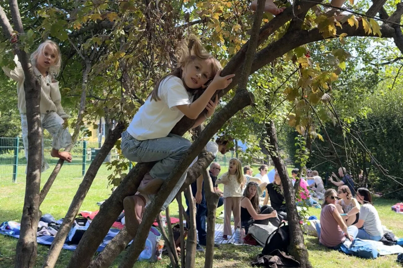Auf dicken Strauchästen sitzen Kinder und schauen in die Kamera. Im Hintergrund ist das ein oder andere Picknick zu sehen.