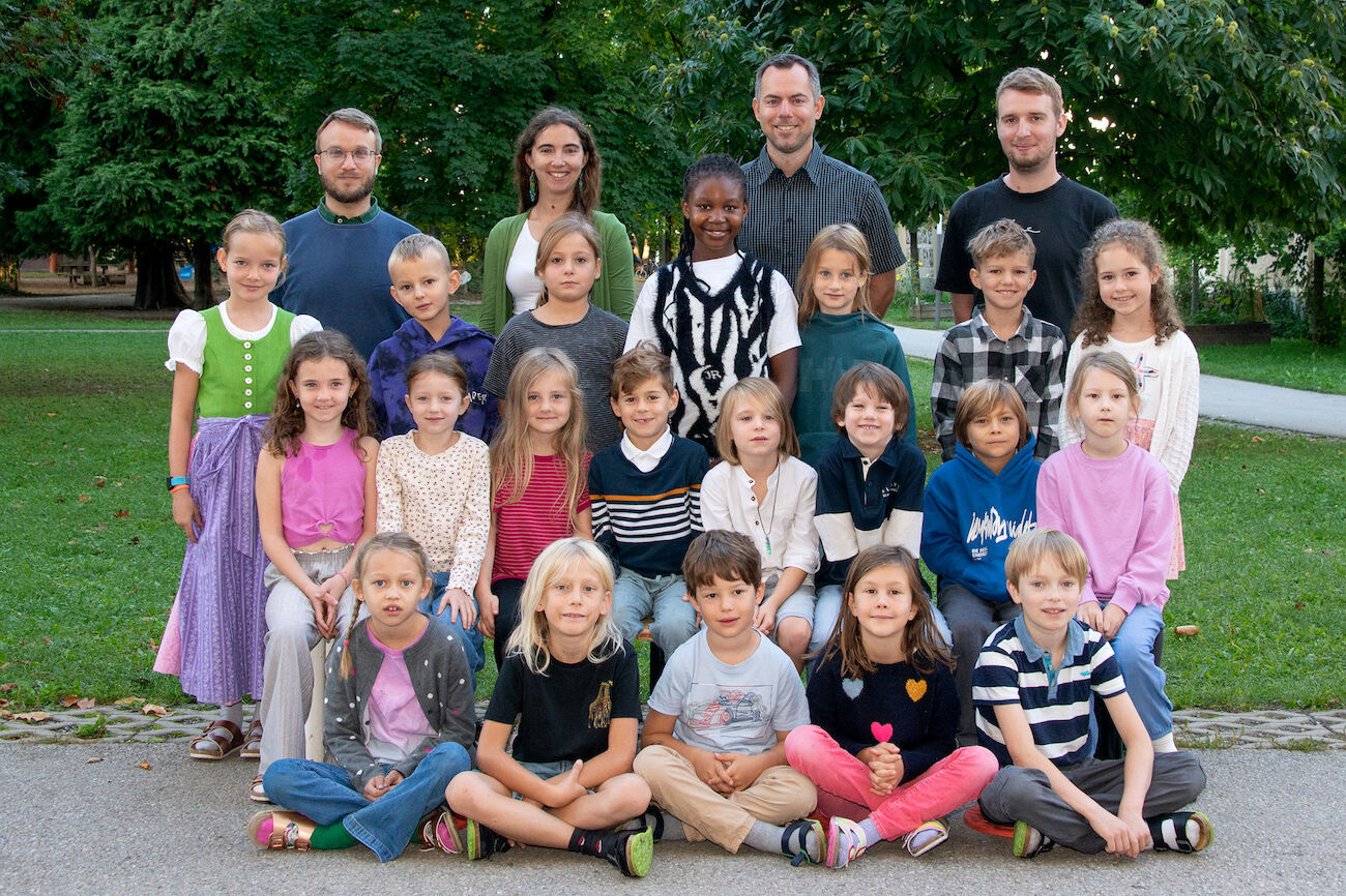 Eine Gruppe von Kindern, zwei Lehrpersonen, ein Nachmittagsbetreuer und ein Schulassistent posieren gemeinsam im Freien für ein Klassenfoto.