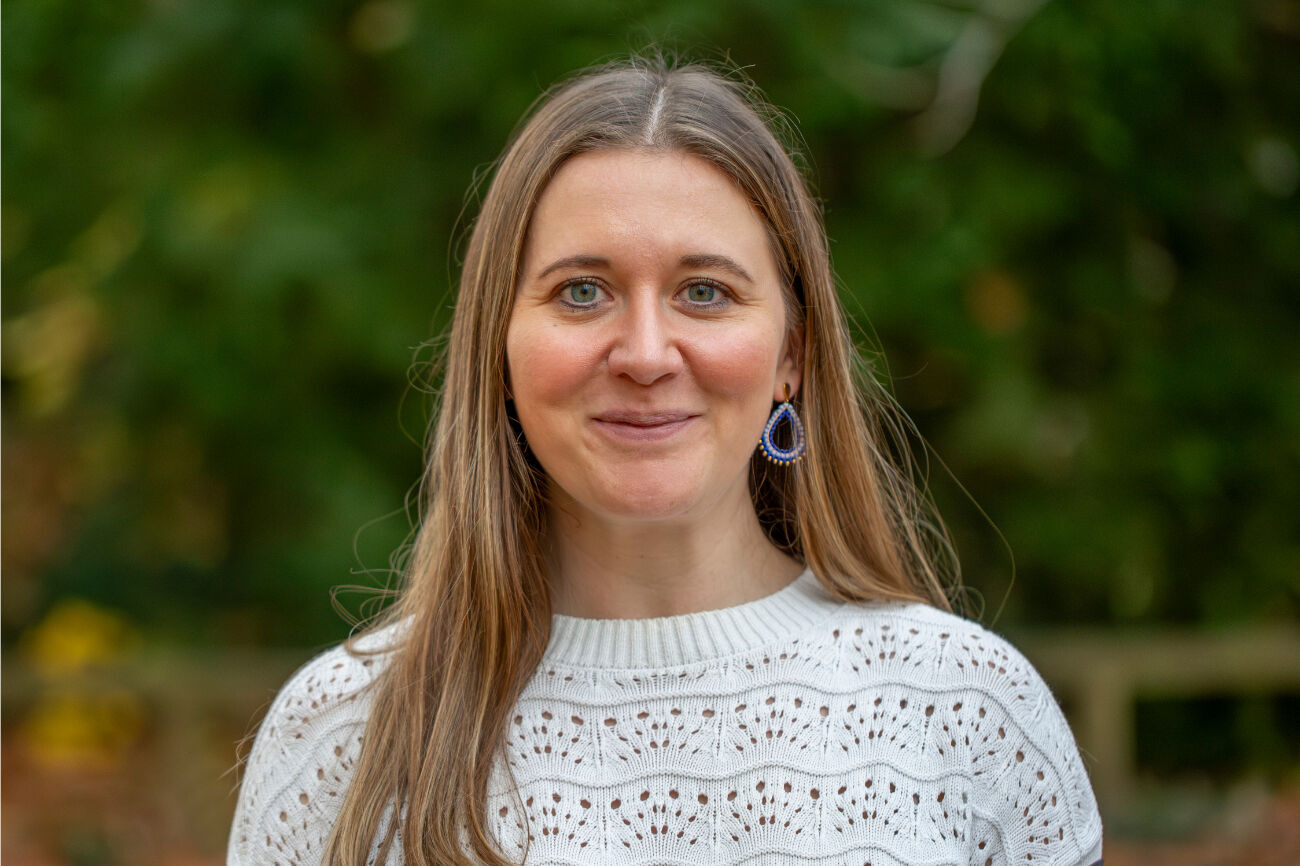 Frau mit langen braunen Haaren, lächelt im Freien und trägt ein rotes Oberteil und eine olivgrüne Jacke.