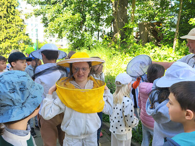 Eine Gruppe von Kindern steht vor einer Wiese zusammen. Ein paar tragen Imkerhüte. Im Hintergrund sieht man leicht versteckt einen Bienenstock und den Imker.