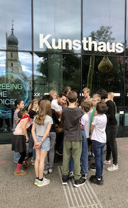 Kindergruppe vor dem Kunsthaus in Graz. In der Glasfassade spiegelt sich der Turm der Franziskanerkirche.