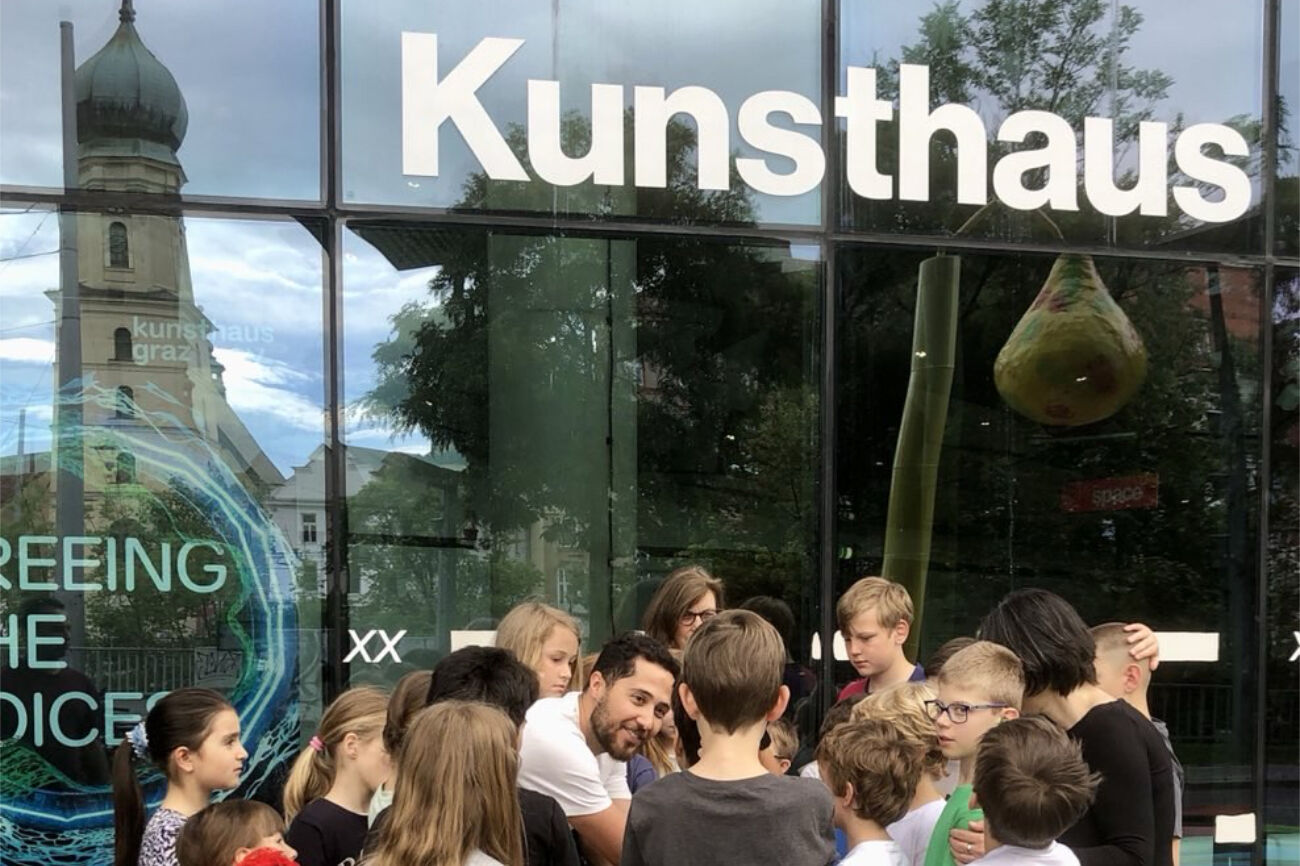 Kindergruppe vor dem Kunsthaus in Graz. In der Glasfassade spiegelt sich der Turm der Franziskanerkirche.