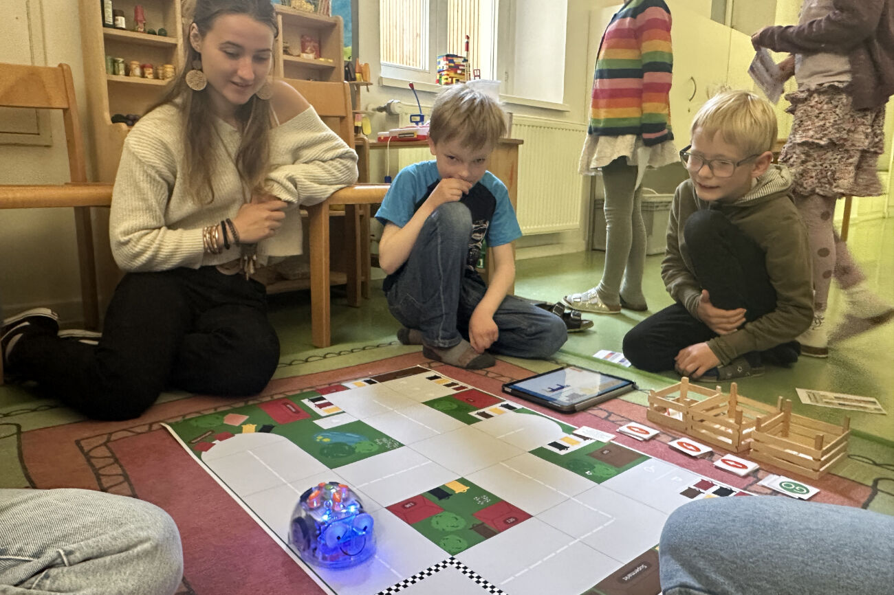 Drei Kinder und eine Frau beobachten, wie sich ein kleiner Roboter auf einem Spielbrett auf dem Boden bewegt.