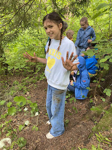 Lächelndes Mädchen mit schmutzigen Händen steht in einem Waldstück, andere Kinder stehen hinter ihr.