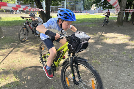 Tief geduckt fahren Kinder auf ihren Fahrrädern unter gespannten Absperrbändern durch.