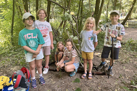 Eine Kindergruppe steht bzw. hockt vor ihrer gebauten kleinen Waldhütte.
