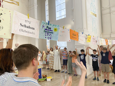 Auf einer Schnur sind Papierblätter mit dem Wort "Frieden" in verschiedenen Sprachen befestigt. Um den Altar stehen dabei die Kinder und Erwachsenen, die gesegnet werden.