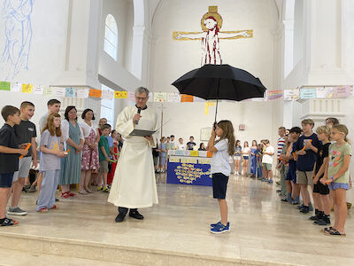Der zukünftige Diakon spricht zum Volk, ein Mädchen mit einem aufgespannten Regenschirm steht ihm zur Seite. Umgeben sind die beiden von den soeben gesegneten Viertklässler*innen und Lehrer*innen.