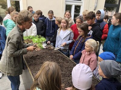 Kinder der PVS und des BischGyms stehen auf der Dachterrasse vor den Hochbeeten und hören einer Professorin zu. Es geht ums Einsetzen von Salatpflanzen.