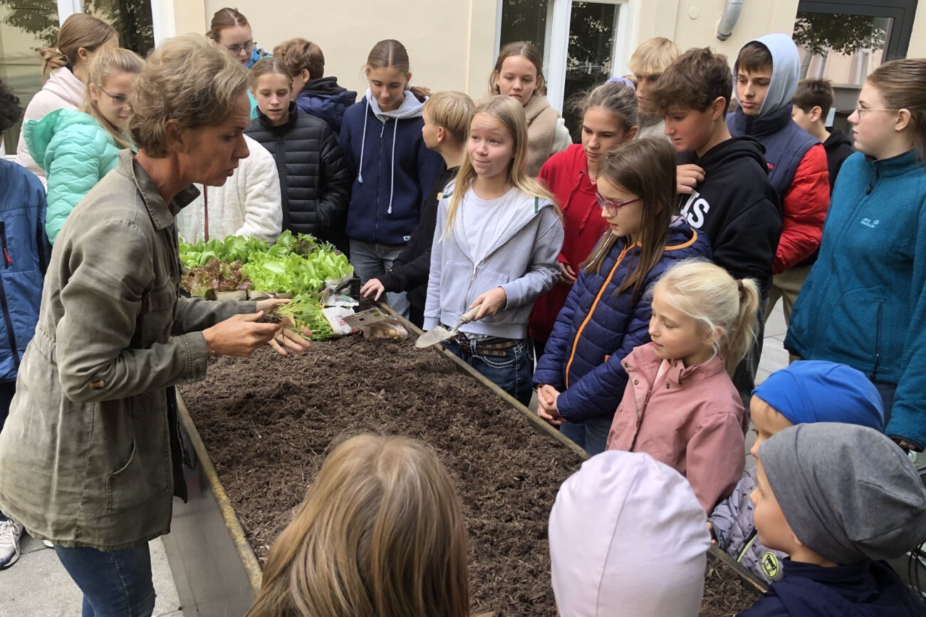 Kinder der PVS und des BischGyms stehen auf der Dachterrasse vor den Hochbeeten und hören einer Professorin zu.