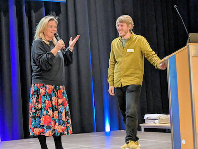 Die Schulleiterin Frau Öttl spricht von der Bühne zum Publikum. Rechts daneben steht ein Kollege von der Nachmittagsbetreuung.