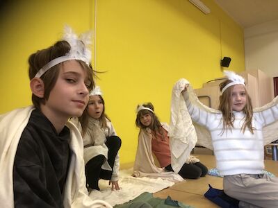 Drei Mädchen und ein Bub beim Theaterspielen auf einer Bühne, verkleidet mit Umhängen und einem Federkopfschmuck.