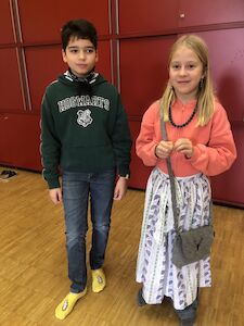 Ein Bub und ein Mädchen präsentieren ihre Verkleidung. Das Mädchen hat neben einem Rock eine Tasche umgehängt.