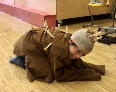 Ein Bub ist als Igel verkleidet und kauert am Boden. Als Stacheln hat er Wäschekluppen auf einem braunen Kostüm festgemacht.