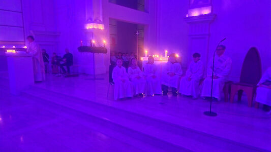 Blick auf den in ein mystisches Lila getauchten Altarraum, wo viele Ministrant*innen sitzen. Im Hintergrund brennen einige Kerzen. Ebenso ist der große hängende Adventkranz zu sehen.