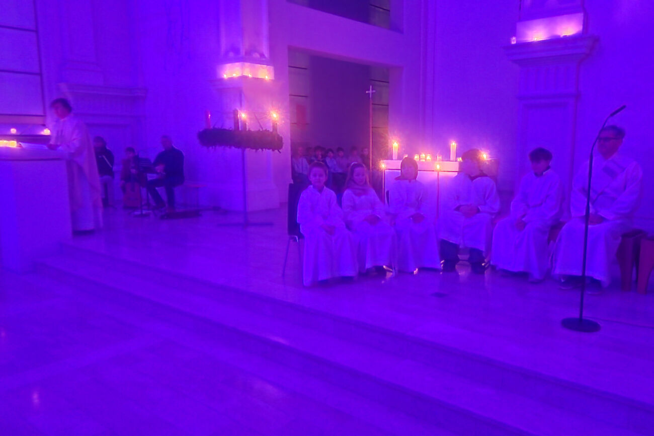 Blick auf den in ein mystisches Lila getauchten Altarraum, wo viele Ministrant*innen sitzen. Im Hintergrund brennen einige Kerzen. Ebenso ist der große hängende Adventkranz zu sehen.