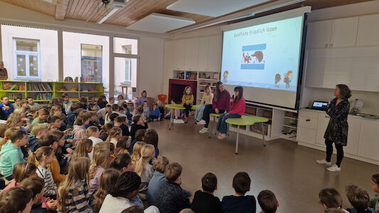 Ein Teil der Delfin-Klasse bei der Podiumsdiskussion im Rahmen der Schulversammlung. Drei Kinder sitzen am Tisch mit einer Lehrerin, im Hintergrund ist ein Bild auf der Leinwand zu sehen.