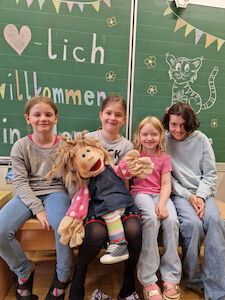Vier Mädchen sitzen mit einer großen Stoffpuppe vor der Kreidetafel, worauf "Herzlich Willkommen" steht.