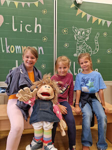 Drei Mädchen sitzen mit einer großen Stoffpuppe vor der Kreidetafel, worauf "Herzlich Willkommen" steht.