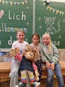 Drei Mädchen sitzen mit einer großen Stoffpuppe vor der Kreidetafel, worauf "Herzlich Willkommen" steht.