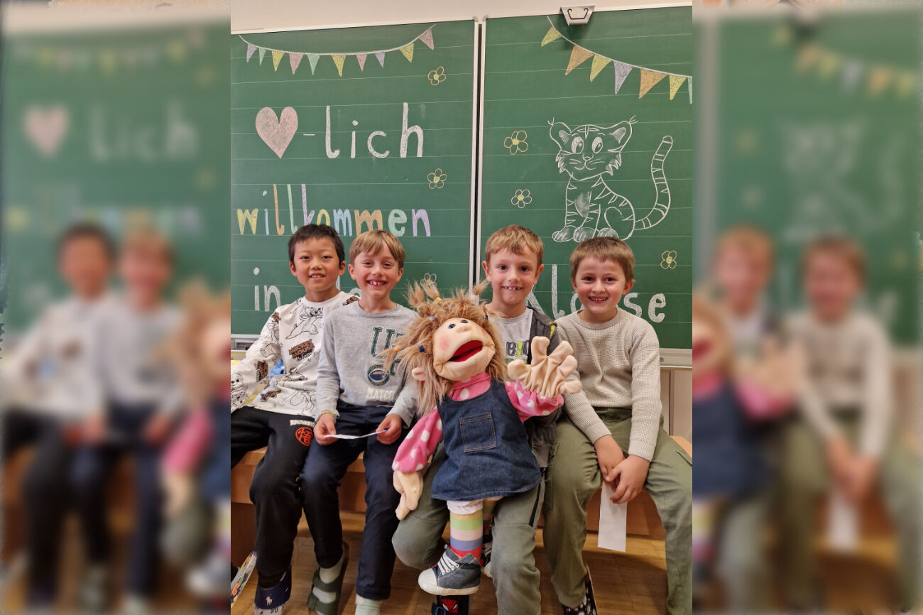 Vier Buben sitzen mit einer großen Stoffpuppe vor der Kreidetafel, worauf "Herzlich Willkommen" steht.