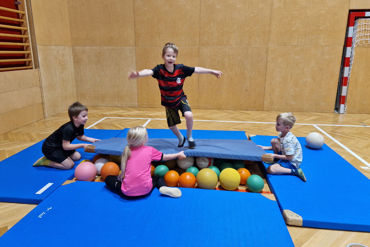 Ein Bub balanciert auf einer Weichmatte, die auf Bällen liegt, umgeben von anderen Weichmatten - gehalten von drei Kindern - damit nichts passiert.