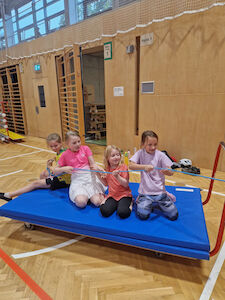 Vier Mädchen ziehen sich mit einem Seil am Mattenwagen sitzend zur Sprossenwand.