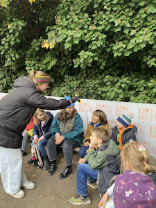 Den Kindern wird im Freien im Kreis sitzend etwas von einer Frau gezeigt.