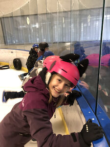Ein Mädchen mit einem rosa Helm steht an der Bande und lächelt.