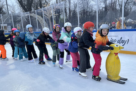 10 Kinder machen am Eislaufplatz einen langen Zug, indem sie sich hintereinander nachfahren und an der Hüfte halten.