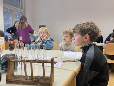 Mehrere Buben sitzen um einen Tisch und beobachten eine Studentin, welche ein Experiment durchführt.