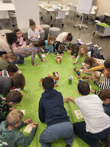 Blick von oben auf die Kinder der Schmetterling-Klasse, die am Boden auf einer grünen Stoffunterlage ihre Bilder zum Gesehenen anfertigen.