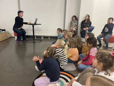 Seitlicher Blick auf die Vortragende und einen Teil der Kinder der Schmetterling-Klasse und ihre Lehrperson im Hintergrund.