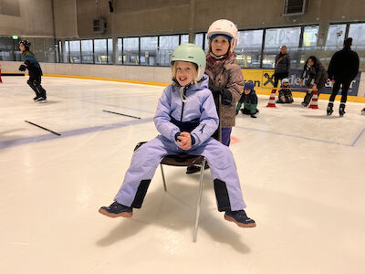 Zwei Kinder haben mit einem Sessel, worauf eines der beiden draufsitzt, großen Spaß auf dem Eis.