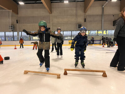 Zwei Buben probieren mit den Eislaufschuhen über ein Holzhindernis am Eis zu springen.