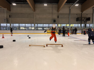 Ein Kind springt gekonnt über ein auf der Eisfläche liegendes Holzhindernis.
