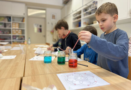 Ein Bub arbeitet mit einer Pipette und drei mit farbigem Wasser gefüllten Gläsern, um ein Muster auf ein laminiertes Blatt zu tropfen.