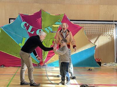 Ein Lehrer unterstützt einen Buben beim Drehen eines Tellers auf einem Holzstäbchen. Clown Jako beobachtet das Tun.
