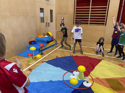 Am Fallschirmtuch befindet sich ein Reifen, worin mehrere Mini-Hütchen abgestellt sind. Drumherum stehen oder bewegen sich einige Kinder.