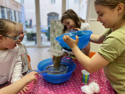 Zwei Mädchen schütten verschmutztes Wasser durch einen selbst gebauten Wasserfilter während andere Kinder aufmerksam das Prozedere beobachten.
