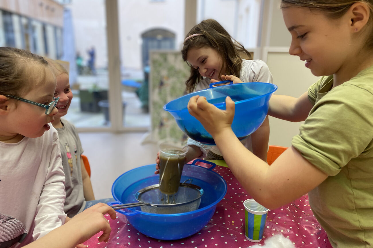 Zwei Mädchen schütten verschmutztes Wasser durch einen selbst gebauten Wasserfilter während andere Kinder aufmerksam das Prozedere beobachten.