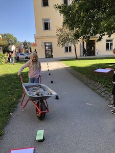 Der Scheibtruhenführerschein wird von einem Mädchen gemacht. Sie muss mit der beladenen Scheibtruhe gerade einen Slalom am Gehweg fahren.